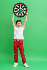 Happy emotional boy of 8 years old, smiling and holding a dart board with a dart in the center...