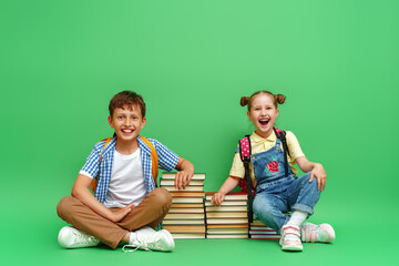 Two happy schoolchildren in loose clothes sit on stacks of school textbooks and reading books, and smile cheerfully against a green background. The concept of children's education. Back to school