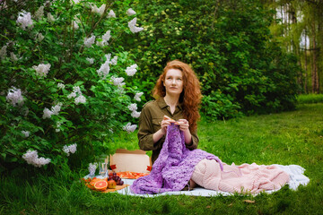 Needlework in the garden. Young woman crocheting sitting on the grass in the park. Picnic and leisure, knitting in nature.