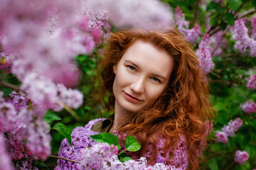 A large portrait of a young beautiful woman with red curly hair near a blooming lilac bush. Copy, empty space for the text.