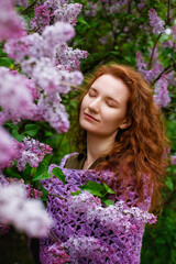 Close up portrait of a young beautiful woman with red curly hair near a blooming lilac bush. The girl dreamily closed her eyes, enjoying the fragrance of flowers. Copy, space for the text. Vertical.