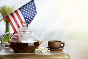 Glass teapot with tea brown cup on a wooden table. American flag in the background. Festive tea party.
