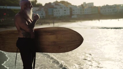 Senior man having fun surfing during sunset time - Retired male training with surfboard on the beach - Elderly healthy people lifestyle and extreme sport concept	