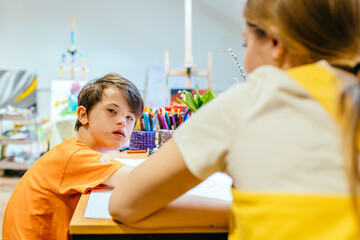 Rear view of nursery teacher sitting with boy child down syndrome looking at camera drawing in the...