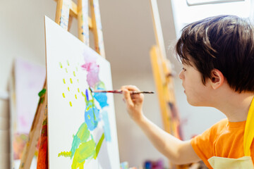 Cute, serious and focused, ten years old boy in orange shirt, apron drawing on canvas on the easel....