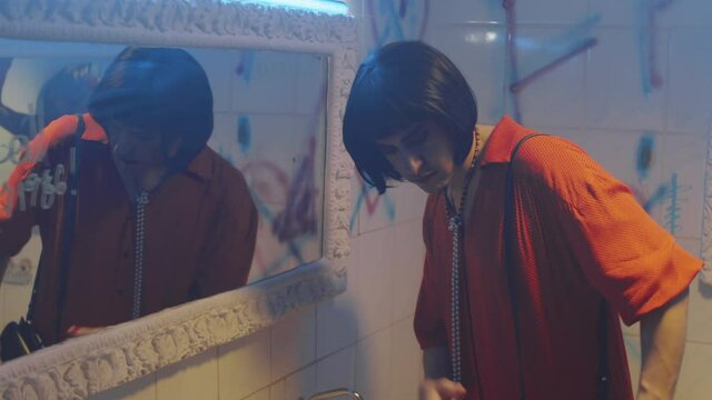 Transgender man in brunette wig and female outfit standing in nightclub restroom, looking at his reflection in mirror and fixing makeup with lipstick