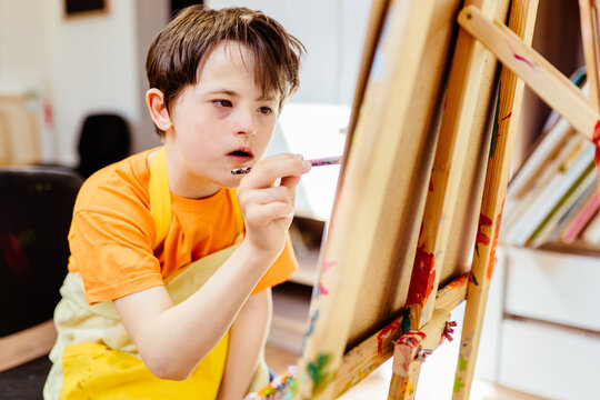 Education and special child concept. Concentrated boy painting on easel. Child with down syndrome holding a brush and drawing at class of studio or home.