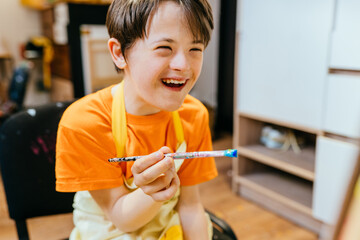 Cute positive happy boy with down syndrome sitting in a chair and drawing with paintbrush indoor.