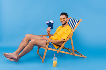 Obraz premium Smiling man holding passports while sitting on deck chair near orange juice on blue background