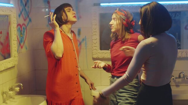 Cheerful transgender man in female dress and wig smoking cigarette and chatting with two girlfriends while standing together in nightclub bathroom