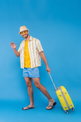 Smiling man in sunglasses waving hand and holding suitcase on blue background