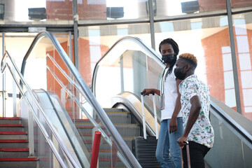 Young african couple wearing protective face masks and carrying baggage climbing the escalator at the subway station.