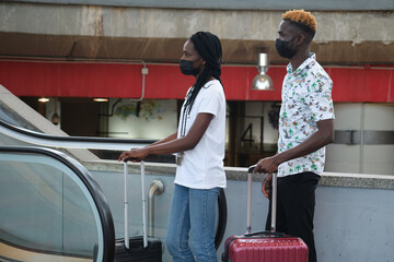 Young african couple wearing protective face masks and carrying luggage climb the escalator at the airport.
