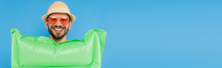 Positive man in straw hat and sunglasses holding inflatable mattress isolated on blue, banner
