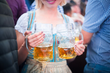 liter beer mugs in the hands of a beautiful smiling girl in a national Bavarian costume dress at Oktoberfest on the terrace with a fresh moody autumn afternoon