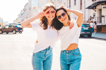 Two young beautiful smiling hipster female in trendy summer white t-shirt clothes and jeans.Sexy carefree women posing on the street background. Positive models having fun, hugging and going crazy