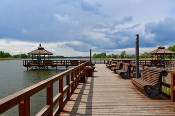 View of a wooden marina, pier or platform with handles and floor made out of boards or logd with a small shack or hut for relaxation located next to river or lake seen before the storm in Poland