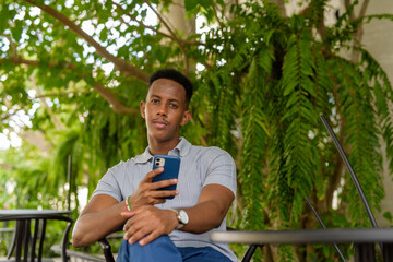 Portrait of young African businessman wearing casual clothes and sitting at coffee shop while holding mobile phone