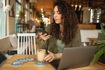 Beautiful young girl working at a coffee shop with a laptop.female freelancer holding a phone connecting to internet via computer, doing online shopping