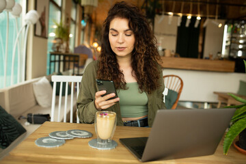 Beautiful young girl working at a coffee shop with a laptop.female freelancer holding a phone connecting to internet via computer, doing online shopping