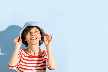 Happy cheerful young woman with short hair, natural makeup over light blue background. Indoor portrait of beautiful brunette young woman wearing blue hat and bright striped clothing	