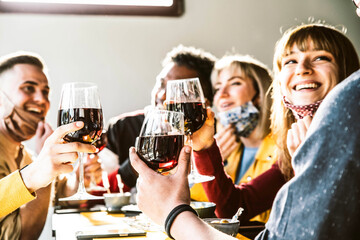 Happy friends wearing protective face masks toasting red wine at bar restaurant - New normal friendship concept with young people having fun at home party - Focus on glasses