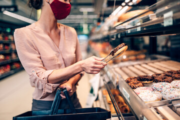 Young woman is buying freshly baked pastry in big modern bakery. She is wearing face protective mask due to Coronavirus epidemic.
