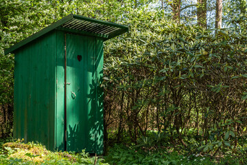 Green timber frame outhouse close to green rhododendron bush. Rustic wooden latrine hided among bushes and blooming trees. Dry outside toilet with heart shape hole in door. Green wood jakes