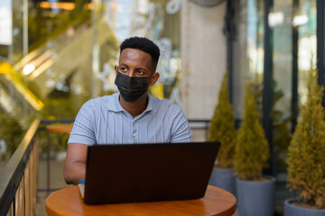 African businessman wearing face mask and social distancing while sitting at coffee shop using laptop computer