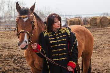 Obraz premium A beautiful woman with long and black hair in a historical hussar costume stands in a field with a horse.