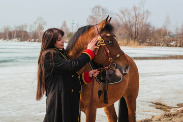 Obraz premium A beautiful woman with long and black hair in a historical hussar costume stands near a river with a horse.