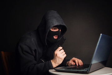 A criminal in a black mask and a hood with a pistol steals money through a laptop. Hacker attacks.