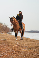 A beautiful woman with long and black hair in a historical hussar costume stands near a river with a horse.