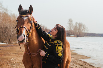 Obraz premium A beautiful woman with long and black hair in a historical hussar costume stands near a river with a horse.