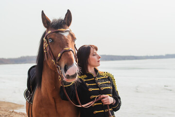 A beautiful woman with long and black hair in a historical hussar costume stands near a river with a horse.