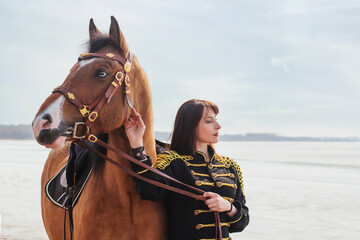 Obraz premium A beautiful woman with long and black hair in a historical hussar costume stands near a river with a horse.