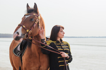Obraz premium A beautiful woman with long and black hair in a historical hussar costume stands near a river with a horse.