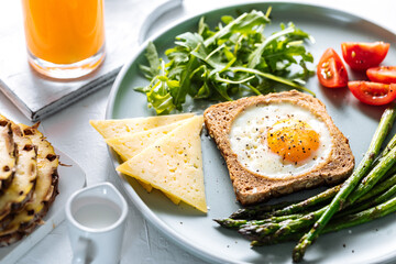 Sunny side up egg in a toast served with cheese, asparagus, rocket and tomatoes on a rustic white background