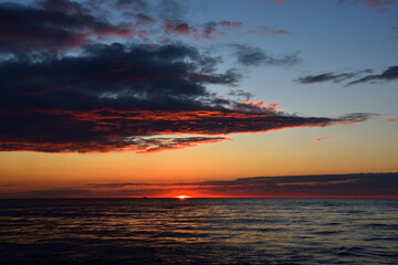 Seascape sunset, protruding above the horizon, the fiery golden edge of the sun paints the dark sea, twilight sky and picturesque clouds in scarlet colors.