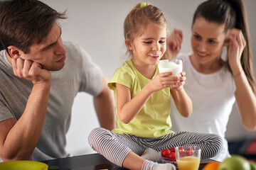 A young happy parents watching their daughter enjoying a yogurt for a breakfast at home. Family,...