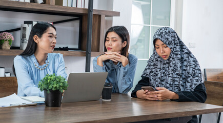 Two Asian women chatting together, and fellow Islamic women wear Hijab holding smartphones, typing messages and online, playing social networks at a company conference table together
