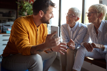 Elderly business women questioning their young male colleague about a smartphone content he is showing to them while they take a break at workplace. Business, office, job