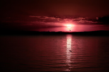 Horizontal dreamy photography of a dark magenta-reddish sunset over calm lake with sunlight reflection on the water