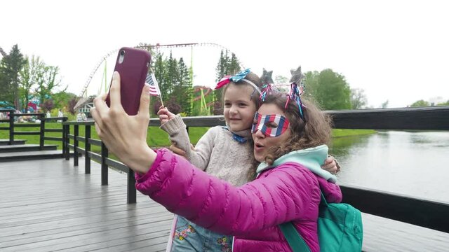 Happy little girl and mother wear symbols usa national flag, smile and talk in video conference by smartphone together, celebrating 4th of July. Activity technology lifestyle mobile phone use concept