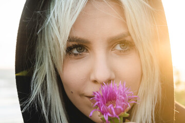 Close up portrait of young nordic blonde woman in hooded jacket while smelling a beautiful flower...