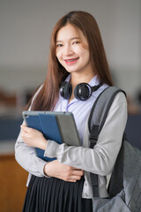 Portrait of young Asian woman student in uniform holding tablet in smart and happy pose in university or college classroom. Youth girl student and tutoring education with technology learning concept.