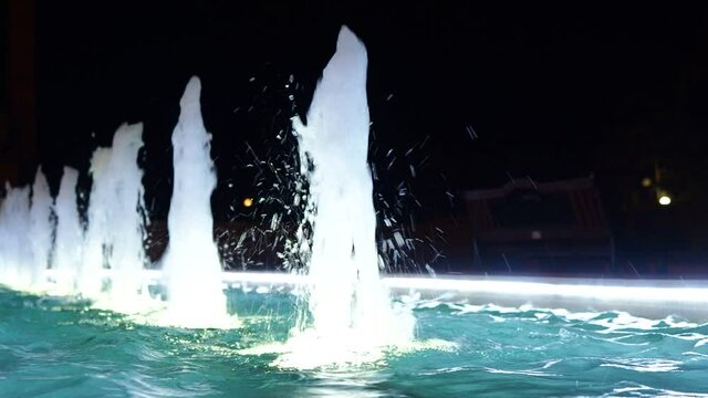 Fountain jets beat in the illuminated turquoise pool in the night park