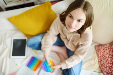 Teenage girl playing with rainbow pop-it fidget toy while studying at home. Teen kid with trendy...
