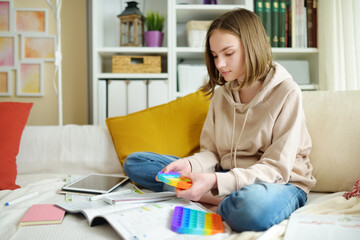 Teenage girl playing with rainbow pop-it fidget toy while studying at home. Teen kid with trendy stress and anxiety relief fidgeting game. - obrazy, fototapety, plakaty