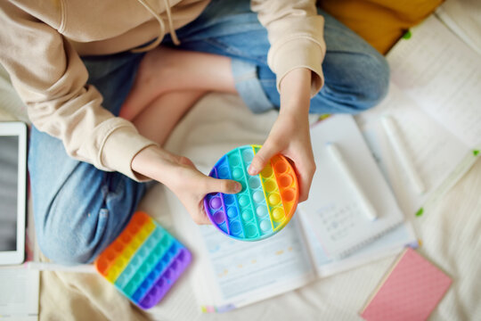 Teenage girl playing with rainbow pop-it fidget toy while studying at home. Teen kid with trendy stress and anxiety relief fidgeting game.